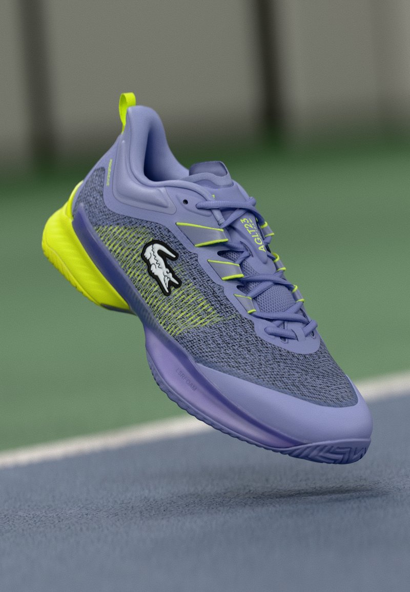 Lacoste sporta apavi lavandas un neona dzeltenā krāsā. Ievērojama elpojoša sieta virsma, teksturēta zole un logotipa akcenti sānos.