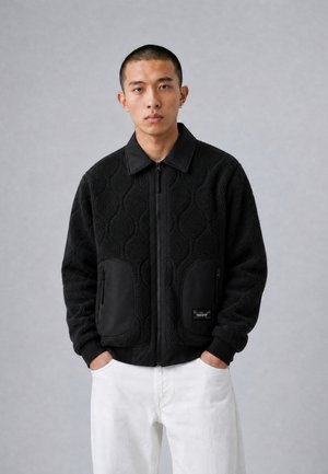Man wearing a black textured zip-up jacket with large pockets and white pants, standing with hands in pockets against a plain background.