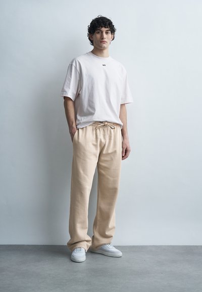 Νεαρός με σγουρά μαλλιά στέκεται φορώντας ένα φαρδύ λευκό μπλουζάκι, μπεζ παντελόνι με κορδόνι και λευκά αθλητικά παπούτσια μπροστά σε έναν απλό γκρι τοίχο.