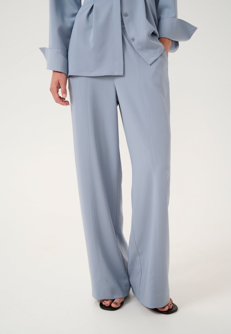 Pantalon large bleu clair associé à une chemise oversize boutonnée assortie, une main dans la poche, talons noirs ouverts visibles.