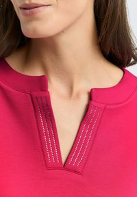 Fuchsia top met een V-hals en een knoopsluiting met vijf verticale rijen kleine zilveren versieringen. Glad stof met een zachte textuur.