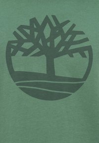 Πράσινο βαμβακερό T-shirt με σκούρο πράσινο, στυλιζαρισμένο λογότυπο δέντρου μέσα σε κυκλικό σχέδιο, πάνω σε ανοιχτό πράσινο φόντο.