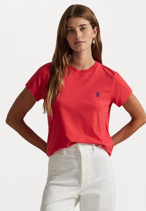 Camiseta de algodón roja con cuello redondo y mangas cortas, con un pequeño logo azul en el pecho. Llevada con pantalones blancos de cintura alta.