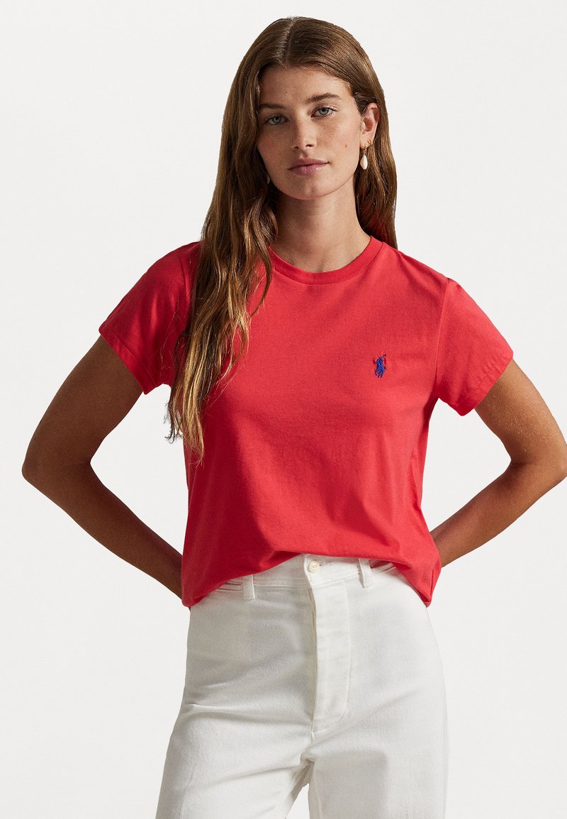 T-shirt in cotone rosso con scollatura a girocollo e maniche corte, con un piccolo logo blu sul petto. Indossato con pantaloni bianchi a vita alta.
