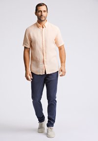 Korte mouw, knoopshirt in licht perzik linnen, gecombineerd met marineblauwe broek en witte sneakers. Model staat rechtop tegen een neutrale achtergrond.