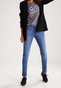 Černý blejzr, šedé tričko s potiskem a textem "navrhl" , modré slim džíny a černé sportovní boty. Jednoduchá a ležérní kombinace oblečení.