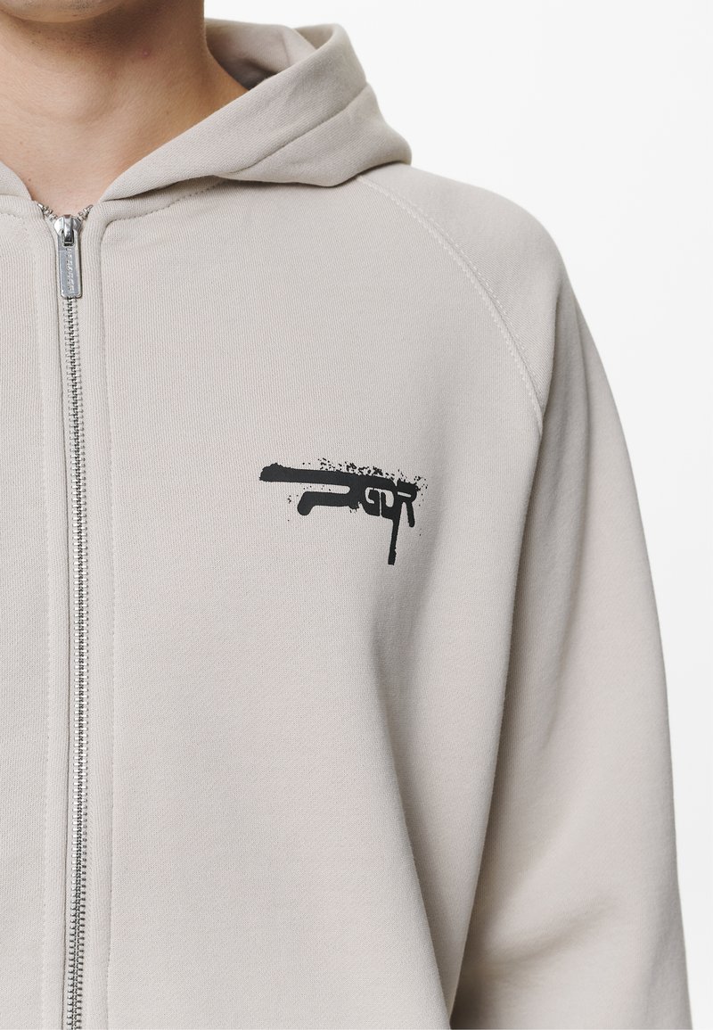 Reißverschluss-Hoodie aus leichtem grauem Baumwollmix mit einem schwarzen grafischen Druck einer stilisierten Feuerwaffe im oberen linken Brustbereich.