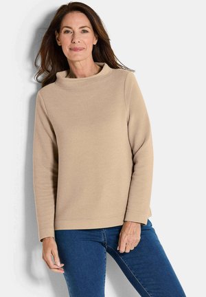 Pull beige à côtes avec un col haut, des manches longues et une coupe décontractée, associé à un jean bleu. La texture est douce et légèrement en relief.