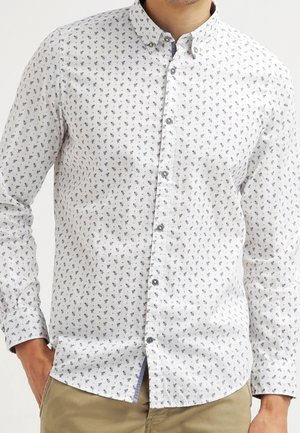 Homme portant une chemise blanche à manches longues avec un petit motif floral foncé et un pantalon beige, les mains dans les poches.