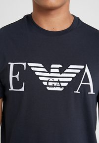 Marinblå bomullströja med vita broderade bokstäver "E", "GA" och "A" med vingliknande streck mellan "GA" och "A".