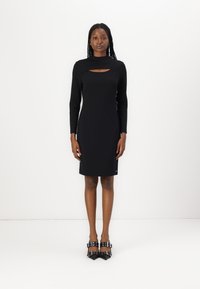Robe noire à manches longues avec une découpe au niveau du col, silhouette ajustée, texture douce et design arrivant aux genoux. Portée avec des talons à boucle noirs.