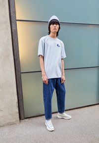 Mladostnik stoji ob zamrznjenem steklu in nosi svetlo sivo kapo, oversize svetlo sivo majico, modre kavbojke in bele visoke superge.