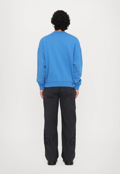 Blaues Baumwoll-Sweatshirt mit Rundhalsausschnitt und gerippten Bündchen, kombiniert mit schwarzen weit geschnittenen Hosen, die ein dekoratives Muster an der Seite haben.