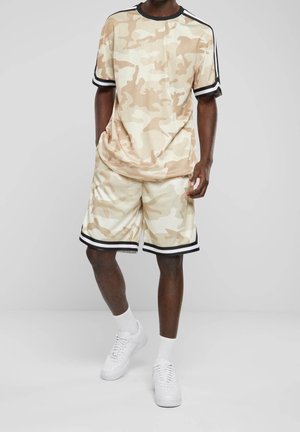 Homme portant une chemise à manches courtes beige et blanche à motif camouflage et un short assorti, avec des chaussettes blanches et des baskets blanches.