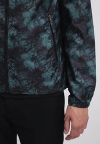 Lekka kurtka na zamek błyskawiczny w ciemnozielonym i czarnym wzorze tie-dye. Posiada elastyczne mankiety oraz oddychającą tkaninę dla większego komfortu.