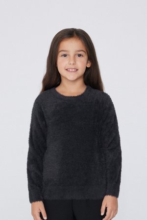 Ragazza giovane con lunghi capelli castani sorride, indossando un maglione grigio scuro peloso e pantaloni neri, in piedi contro uno sfondo chiaro e uniforme.