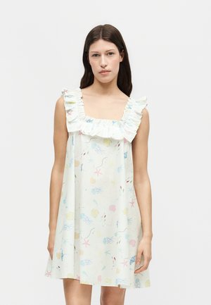 Jeune femme portant une robe blanche sans manches avec un col carré à volants et un imprimé pastel de créatures marines sur fond uni.