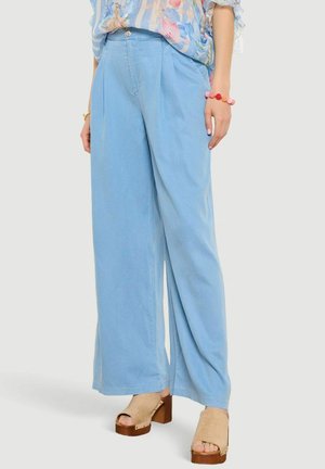 Žena v svetlomodrých nohaviciach s širokými nohavicami, kvetinovej blúzke, ružovom náramku s korálikmi a béžových platformových sandáloch s otvorenou špičkou, stojaca na jednofarebnom pozadí.