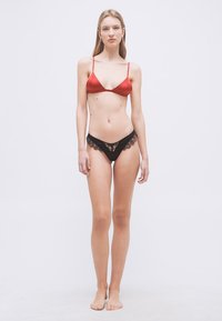 Γυναίκα όρθια, ξυπόλυτη, φορώντας κόκκινο σουτιέν και μαύρο εσώρουχο με δαντέλα, σε απλό λευκό φόντο.