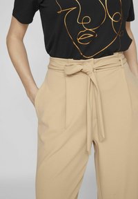 Beige hooggerande broek met een strikdetail bij de taille, gecombineerd met een zwarte t-shirt met een abstract ontwerp van een gouden gezichtsomtrek.