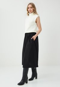 Donna in piedi, che indossa un maglione bianco senza maniche con colletto alto, una gonna midi nera con tasche e stivali alti neri con tacco, su uno sfondo neutro.