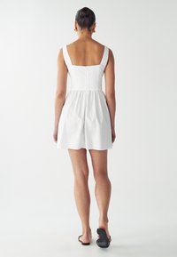 Vestido corto blanco con espalda cuadrada, tirantes anchos, cintura fruncida y tejido suave y ligero. Llevado con sandalias negras.