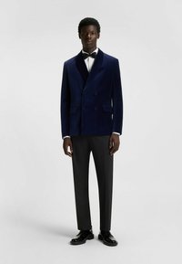 Marineblauw fluwelen dubbelzijdig jasje met reverskraag, zwarte formele broek en glanzende zwarte schoenen, tegen een neutrale achtergrond.