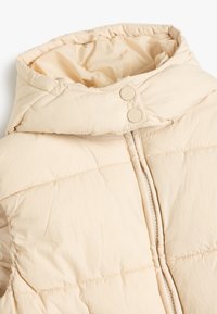 Beige dunjakke med høj krave, to trykknapper og en central lynlås. Stoffet har en let glans og quiltet tekstur.