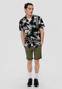 Korte mouw zwarte shirt met een wit bloemenpatroon, gecombineerd met olijfgroene shorts met opgerolde zoom; het model draagt witte sokken en zwarte sneakers.