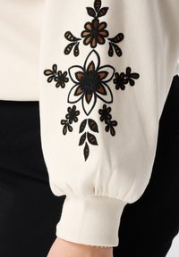 Crèmekleurige top met zwart en bruin bloemenborduursel op de mouw; voorzien van een geplooide manchet en een zachte textuur.