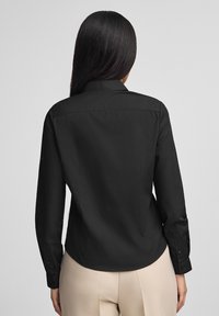 Maglietta nera a maniche lunghe con colletto strutturato, texture liscia e polsini con bottoni. Presenta una vestibilità sartoriale e una sottile cucitura sul retro.