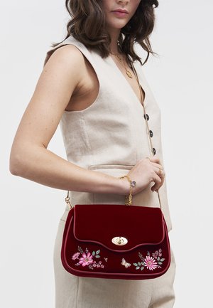 Femme portant un gilet sans manches beige et un pantalon, tenant un sac à main en velours rouge avec broderie florale et fermoir doré.