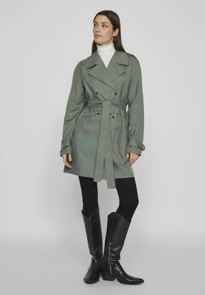 Olijfgroene trenchcoat met dubbele knopen, tailleband en gestructureerde kraag, gecombineerd met zwarte leggings en hoge zwarte laarzen.