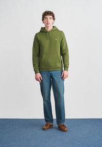 Πράσινο φούτερ με κουκούλα και τσέπη καγκουρό, συνδυασμένο με μπλε τζιν και καφέ παπούτσια. Το μοντέλο στέκεται μπροστά σε απλό φόντο.