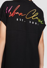 Chemise noire à manches courtes avec manches retroussées arborant un texte coloré "Urban Classic EST. 2005" sur le haut du dos.