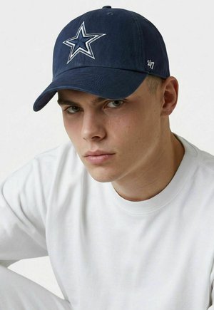 Young man wearing a navy baseball cap with a white star emblem and a white crew-neck sweatshirt, looking directly ahead.
