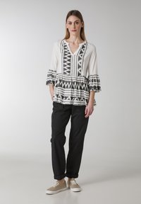 Vit topp med svarta geometriska mönster, V-ringning och trumpetärmar, tillsammans med svarta byxor och bruna sneakers. Slät tyg, avslappnad design.