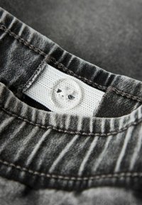 Grå denimstoff med kontrasterende søm; har en hvit, ribbet etikett med en klar knapp og et dekorativt trådmønster.