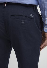 Pantaloni di cotone blu navy con una tasca posteriore dotata di chiusura a bottone e un'etichetta. Il tessuto ha un aspetto texture.