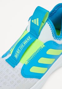 Pantof cu sport cu un superior din plasă albă, accente albastre și o curea verde neon etichetată „ÎNVINGE CĂLDURA”. Talpă netedă cu detalii texturate.
