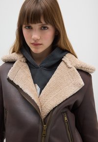 Giacca in pelle marrone con colletto in finta pelliccia color crema, davanti con zip e tasche laterali; indossata sopra una felpa scura con cappuccio.