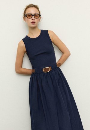 Žena v tmavomodrých šatách bez rukávov, s hnedým opaskom a jantárovo sfarbenými slnečnými okuliarmi v tvare mačacích očí, stojaca pred jednoduchou svetlou stenou.