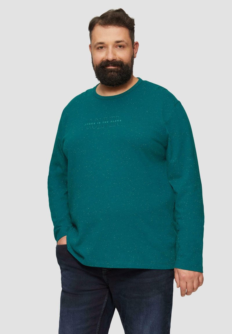 Türkisfarbiges Langarmshirt aus strukturiertem Stoff, mit rundem Halsausschnitt und geprägt textlichem Design auf der Brust. Trägt man mit dunklen Jeans.