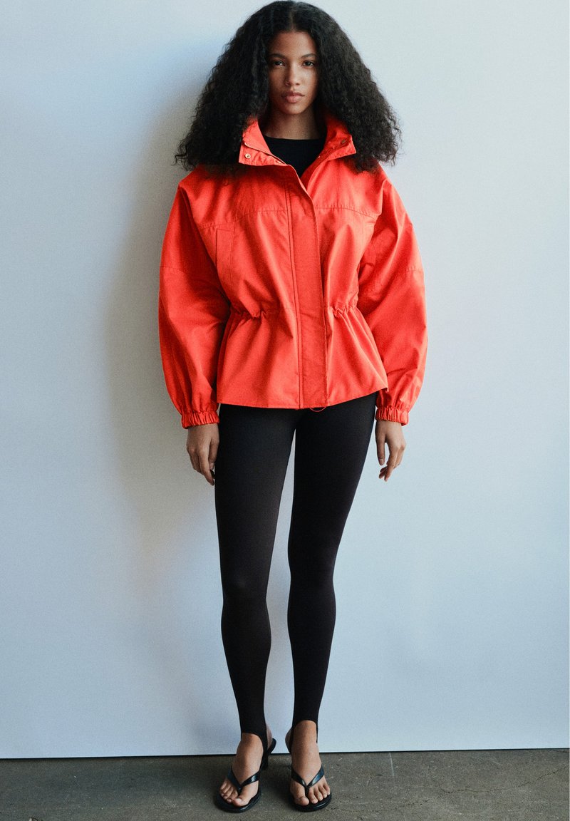 Femme aux cheveux bouclés portant une veste orange, des leggings noirs et des sandales noires, debout contre un mur gris clair.