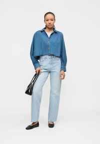 Γυναίκα με πλεξούδες, φοράει cropped τζιν μπουφάν, ανοιχτόχρωμα μπλε τζιν, μαύρα μπαλαρίνες και κρατά μια μαύρη τσάντα χειρός.