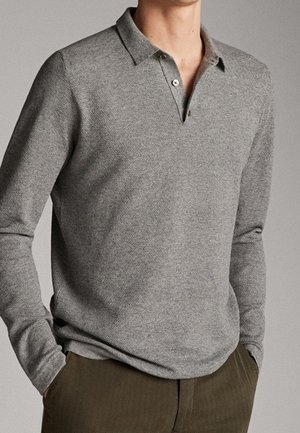 Homme portant un polo gris à manches longues avec deux boutons, les mains dans les poches, et un pantalon vert olive sur un fond neutre.