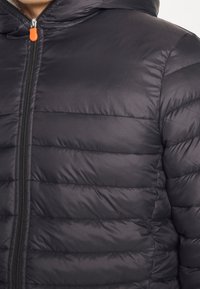 Giacca a vento nera con motivo a cuciture orizzontali, chiusura a zip frontale completa, cappuccio e dettaglio arancione sul tirazip. Finitura in tessuto liscia e lucida.