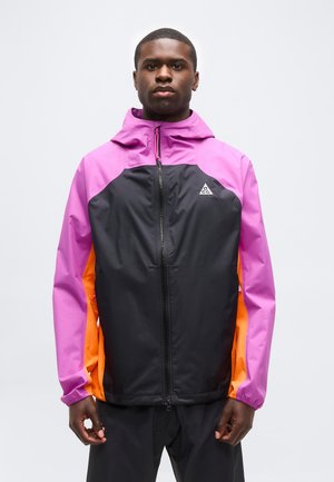 Mies, jolla on musta, pinkki ja oranssi vetoketjullinen huppari, seisoo vaaleaa taustaa vasten kasvot eteenpäin.