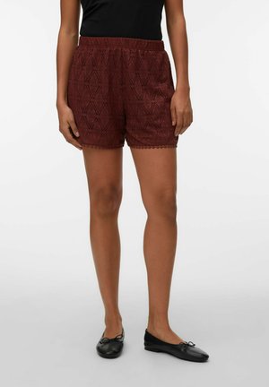 Kobieta ubrana w bordowe koronkowe szorty, czarny top i czarne baleriny, stoi na tle jednolitego jasnego tła.