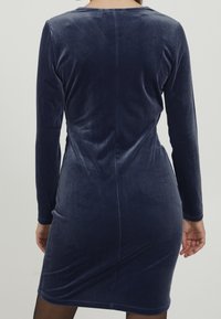 Knieblaues Velvetkleid mit langen Ärmeln und taillierter Form, das eine glatte Textur und eine Nahtdetail entlang des Rückens aufweist.
